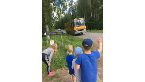 Neljä lasta odottaa myymäläautoa risteyksessä ja yksi lapsista vilkuttaa kuljettajalle kädellään.