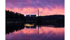 Perävaunillinen rekka-auto ajaa purppuran ja oranssin sävyttämän taivaan alla sillalla. Joki heijastaa näkymän kauniisti.
