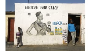 Nuori poika ja aikuinen mies hengailevat parturin ulkopuolella. Valkoisella seinällä on iso mainosmaalaus, tekstinä yläpuolella "African Hair salon" sekä tekstit Black Like Me.
