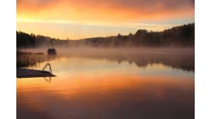 Auringonnousun oranssiksi värjäämä järven huuruava lahti.