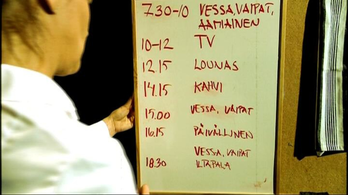 Hoitaja etualalla pitelee taulua, johon on kirjoittettu päivän aikataulu (vessa, vaipat ja aamiainen, TV, lounas, kahvi, vessa ja vaipat, päivällinen, vessa, vaipat ja iltapala).