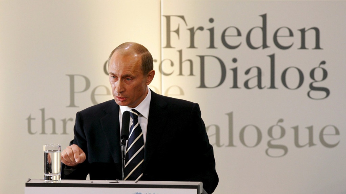 Vladimir Putin puhuu puhujankorokkeen takaa. Hänellä on musta puku ja musta-valkoraitainen kravatti. Korokkeella on mikrofoni ja vesilasi. Putin korostaa juuri sanojaan osoittamalla oikean kätensä etusormella korokkeeseen. Taustalla lukee Frieden durch Dialog eli rauhaa vuoropuhelun kautta.