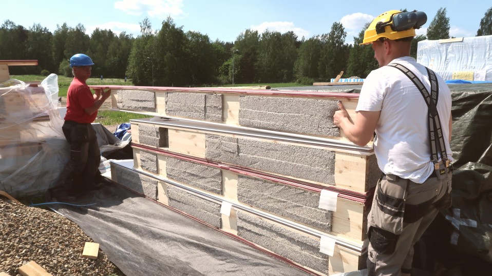 Kaksi miestä nostaa pientä taloelementtiä työmaalla.