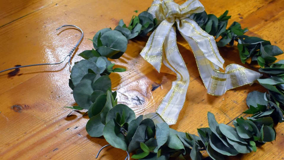 Kuva valmiista kranssista, joka on tehty eukalyptuksen oksista. Kranssin päälle on liimattu jouluinen kultaa kimaltava rusetti.