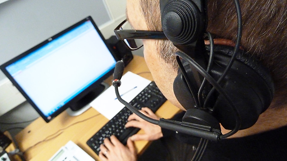 Mies istuu tietokoneen äärellä headset-luurit päässään