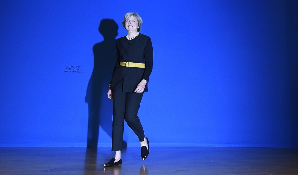 Tummaan housupukuun, leveään keltaiseen vyöhön ja kiiltonahkakenkiin pukeutunut Theresa May kävelee lavalle.
