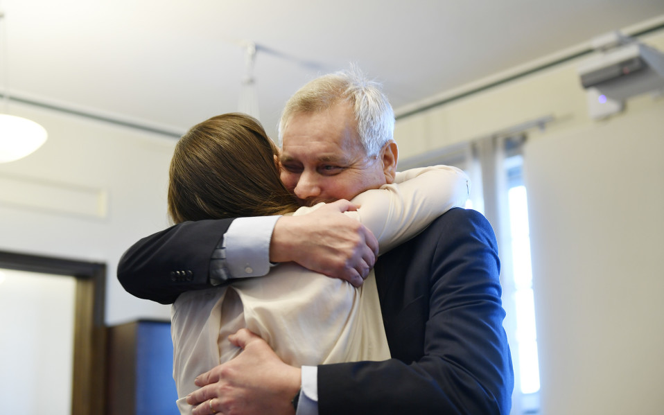 Antti Rinne, en äldre man med grått hår i kostym omfamnar Sanna Marin. Kvinnan har en ljus tröja och brunt hår.