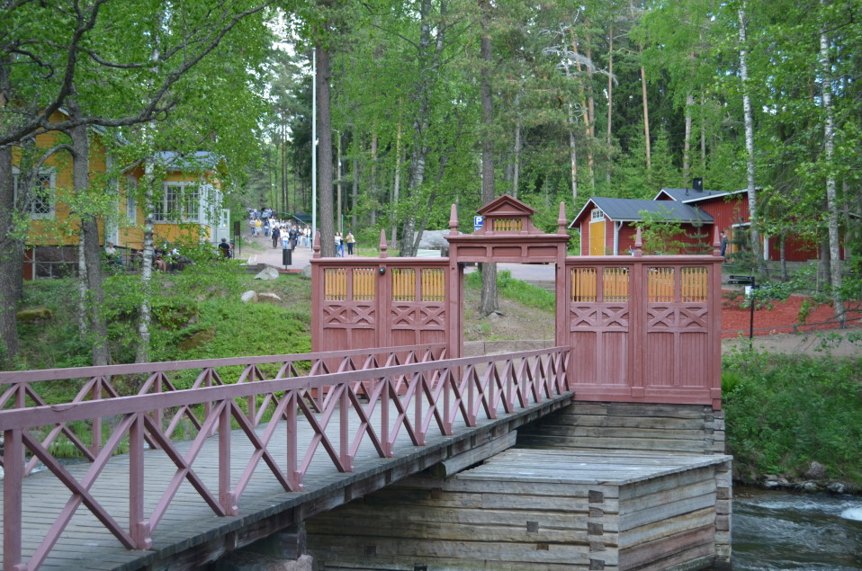 Silta, sillan päässä punertava Langinkosken keisarillisen kalastusmajan portti. Suuri joukko ihmisiä kävelemässä mäkeä alas kohti museon sisäänkäyntiä.