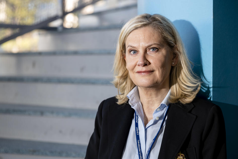Vaaleahiuksinen nainen katsoo suoraan kameraan. Kauluspaita ja siisti olemus. Takana seinä ja portaat.