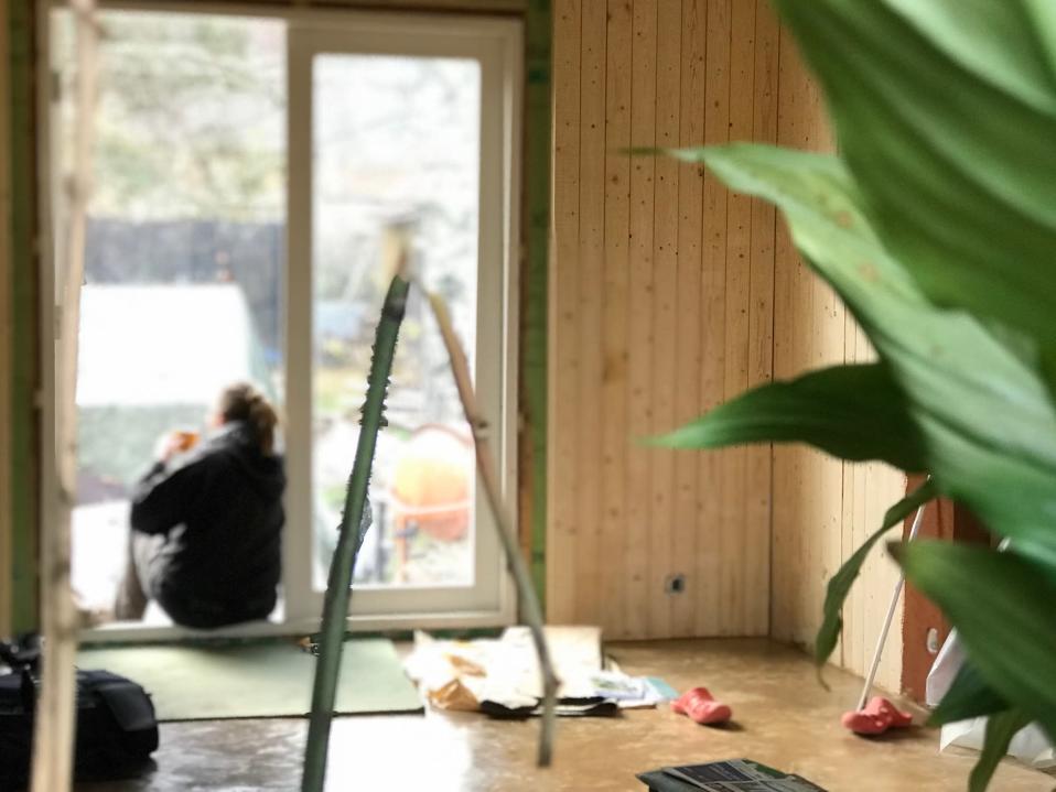 Taustalla nainen istuu selin kuistin oviaukossa, etualalla viherkasvi