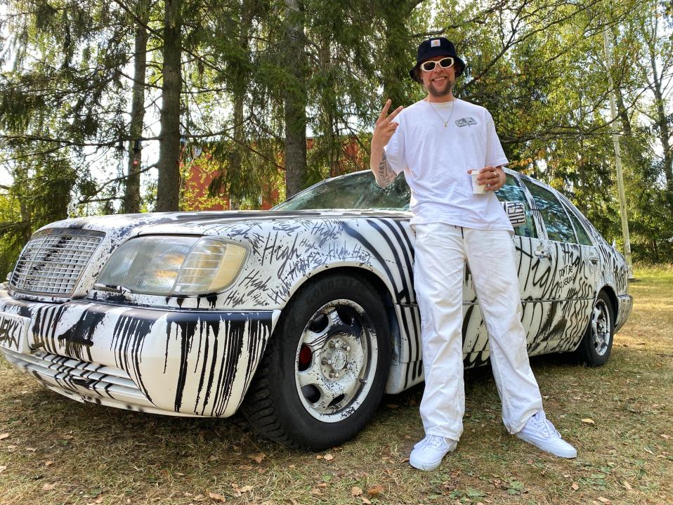 Taiteilija Sheikki keravalaisen purkutaidetalon pihalla oman Mercedes-Benz-autonsa edessä.