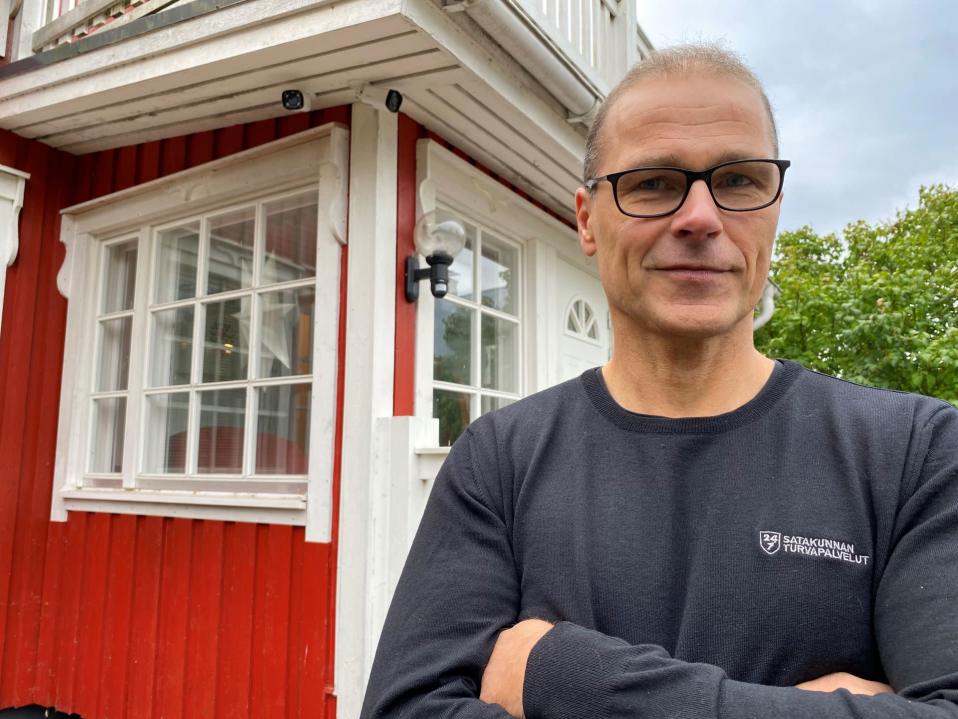 Hannu Tarkkio seisoo punaisen omakotitalon portailla. Hänen olan takaa näkyy kaksi valvontakameraa