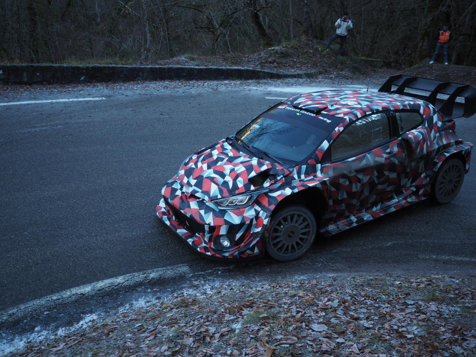 Kalle Rovanperä testaamassa Toyotan uutta hybridiautoa Ranskassa. Rovanperä kurvaa parhaillaan vasemmalle kääntyvään kurviin. Taustalla muutama katsoja seuraamassa suomalaisen testiajoa.