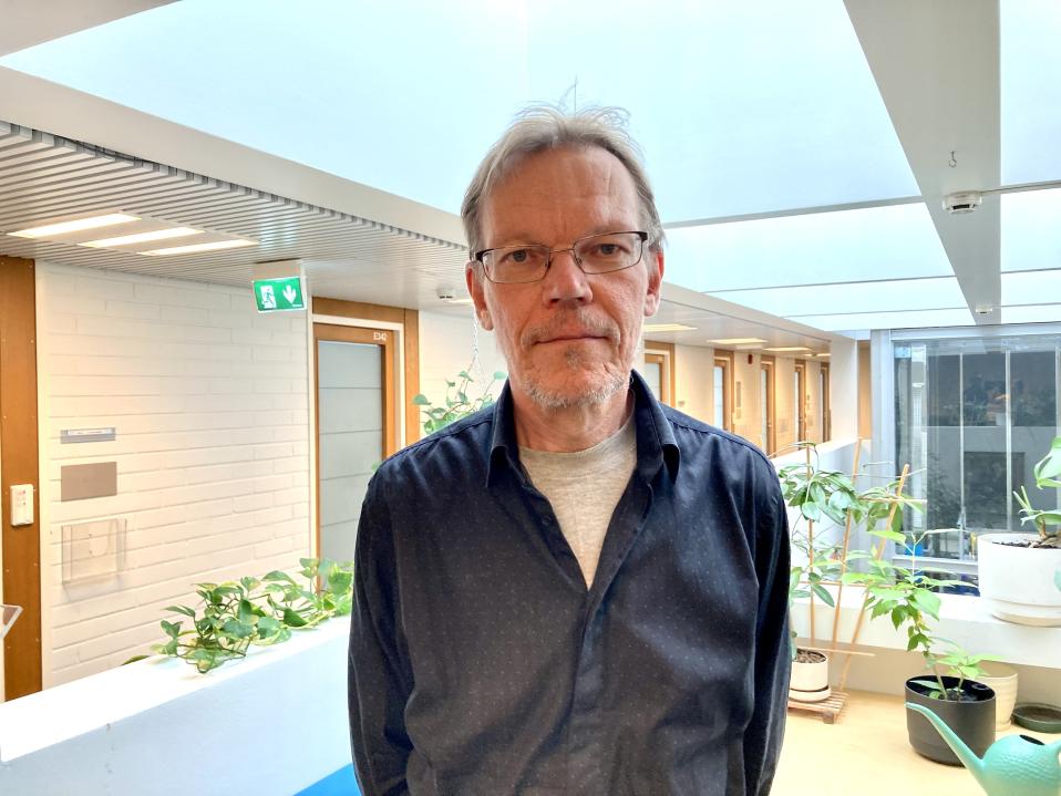 Silmälasipäinen mies seisoo toimistossa, taustalla on käytäväovia ja huonekasveja.