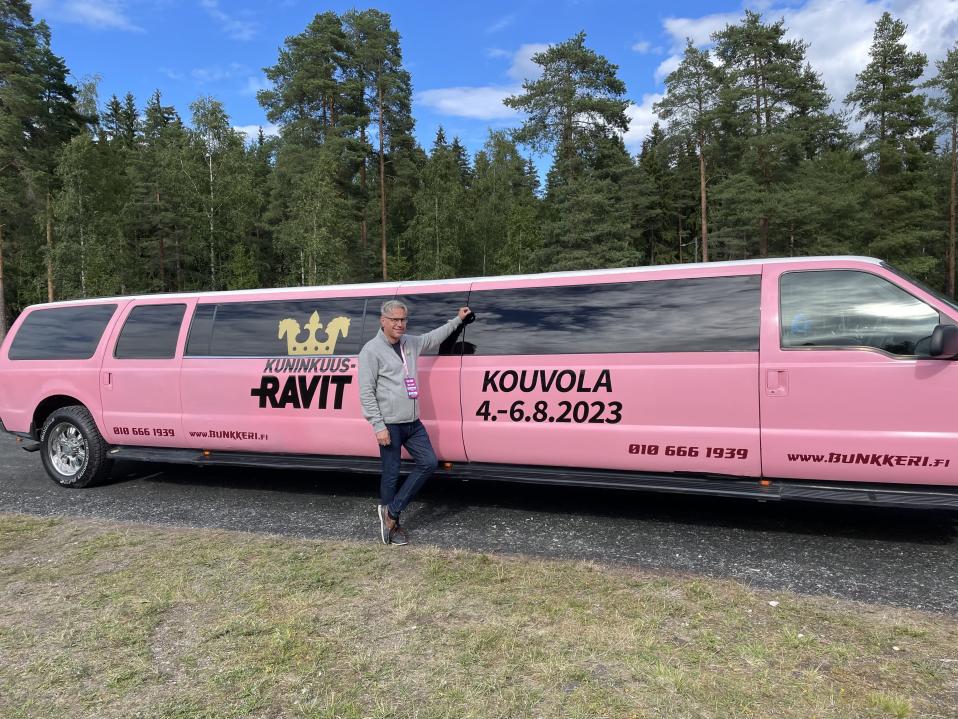 Mies seisoo vaaleanpunaisen limusiinin edessä ja näyttää energiseltä. Autossa teksti