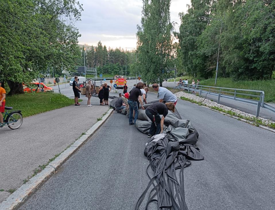 Kesäillassa ihmisiä kerääntyneenä kasaamaan joukolla kuumailmapalloa pois autotieltä. Paikalla pallon kimpussa 10-20 ihmistä, kauempana näkyy pelastusajoneuvoja.