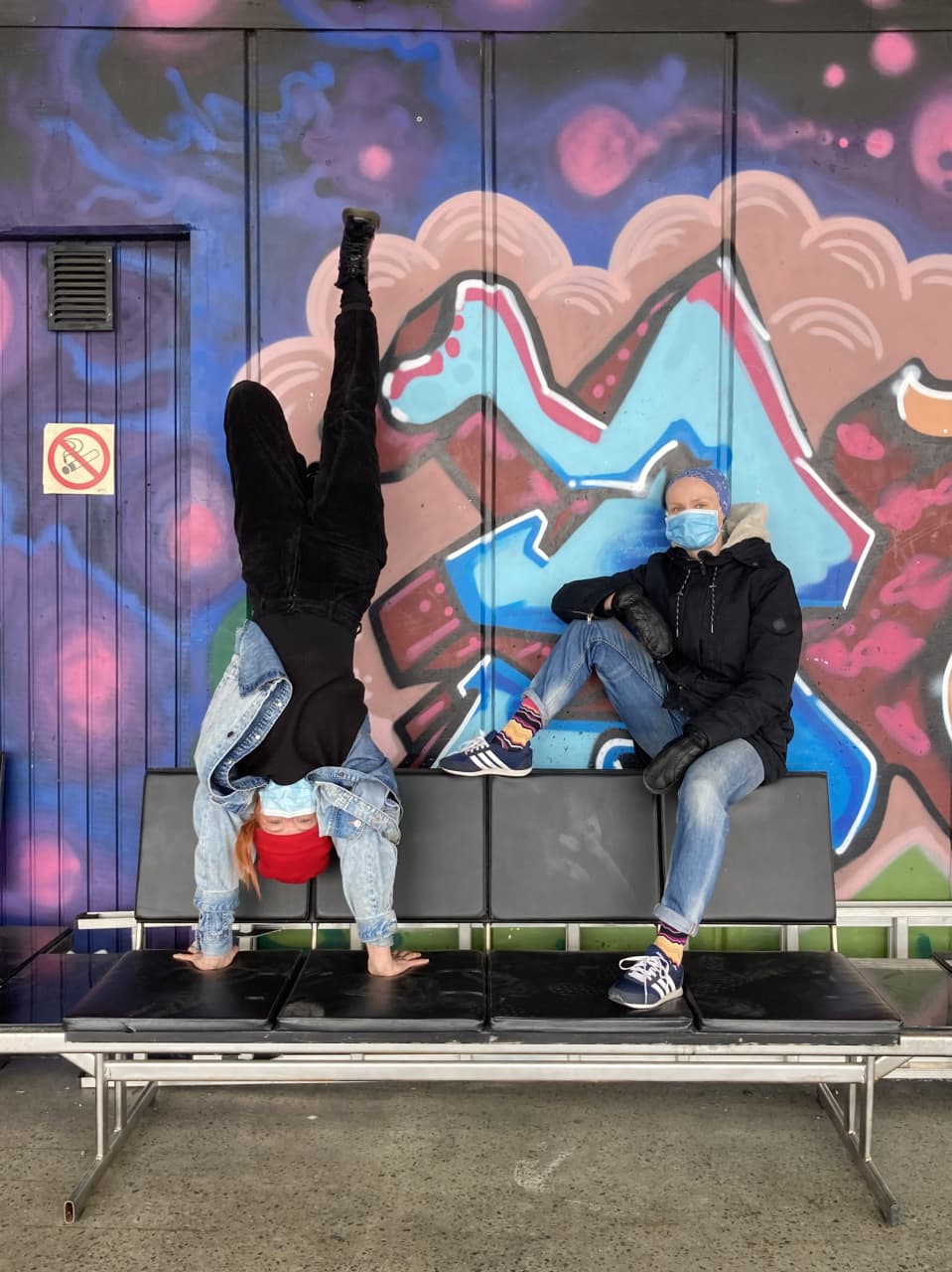 Valpuri Kaarninen istuu penkillä ja Rosa-Maria Autio seisoo käsillään hänen vieressään. Taustalla on sirkuksen iloisenvärinen graffitiseinä.