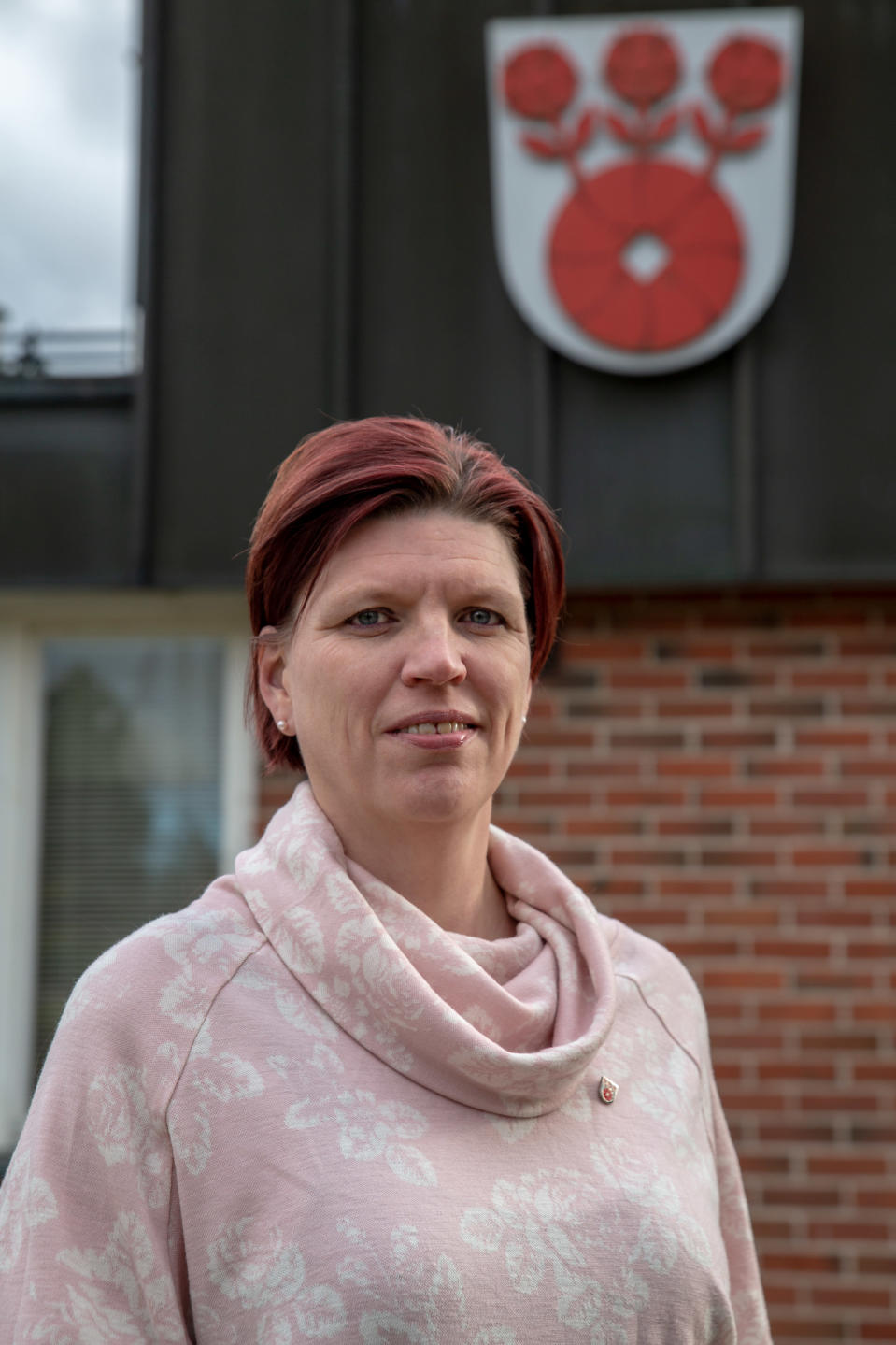 Valtuuston puheenjohtaja Sanna Kurki seisoo Askolan kunnantalon edessä ja katsoo kameraan. Kunnan vaakuna näkyy taustalla kunnantalon seinällä.