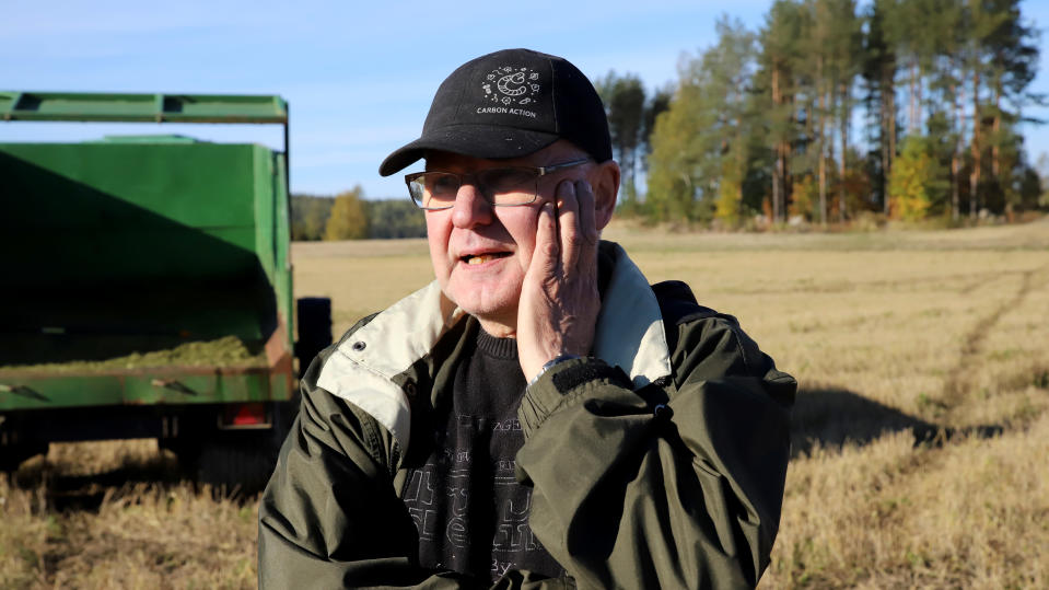 Maanviljelijä Ilpo Markkola lippahattu päässä, toinen käsi poskella seisoo pellolla, takana puimuri.