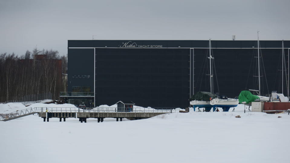 Veneiden säilytyshalli Yacht Store Kotkassa ulkoa päin kuvattuna. Tummansininen rakennus, pihalla pieni mökki ja auto.