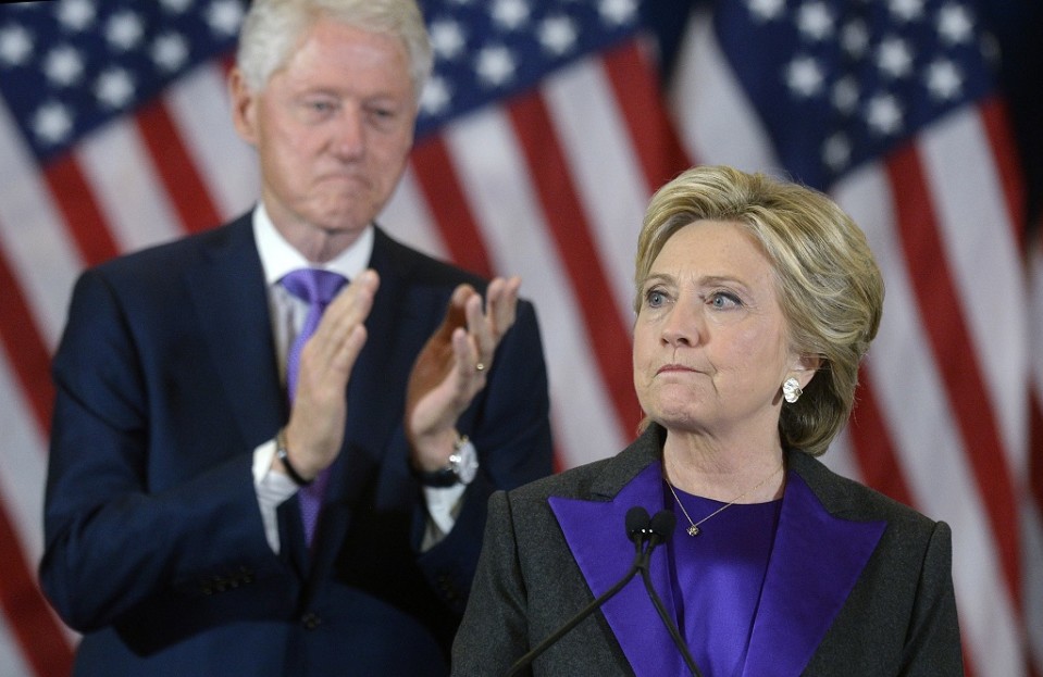 Hillary Clinton katsoo kuvan vasempaan laitaan vakavana. Taustalla taputtaa Bill Clinton. Taustalla on myös Yhdysvaltain lippuja.