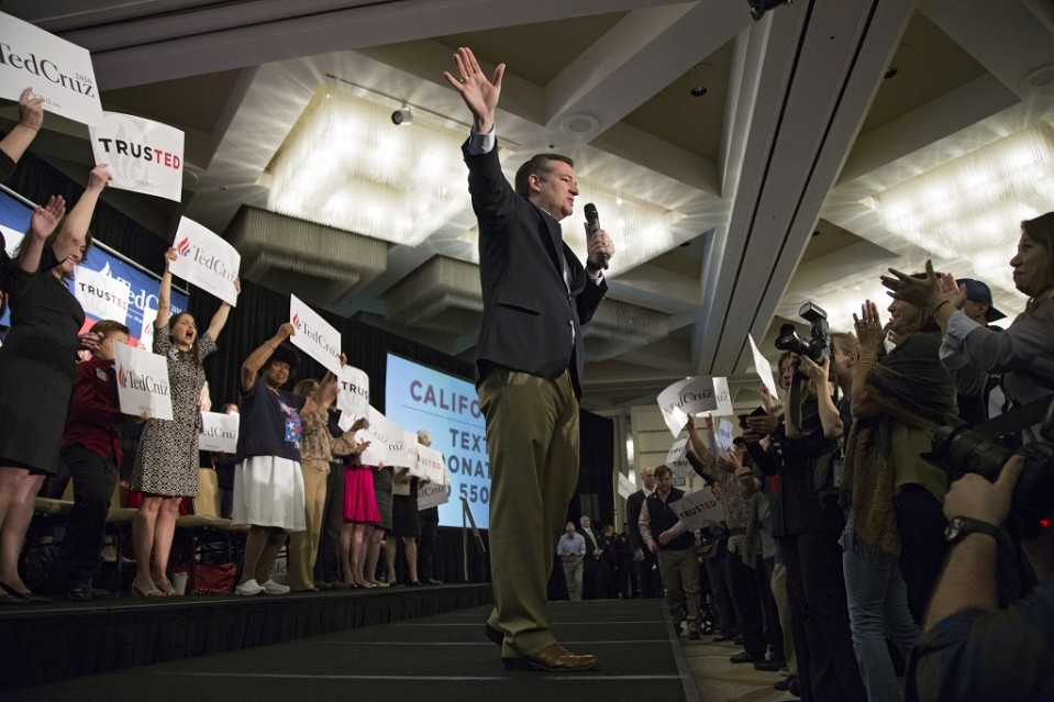 Ted Cruz puhuu vasemmassa kädessään olevaan mikrofoniin ja viittilöi oikealla kädellään jonnekin kauas. Kuva on otettu alaviistosta. Cruzin takana seissoo naisia, jotka pitävät käsissään hänen kampanjajulisteitaan.