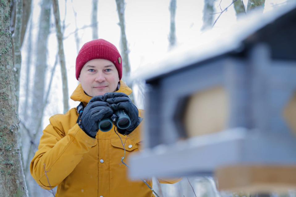Ossi Nokelainen kiikarit kädessä talvisessa metsässä, etualalla lintulauta.