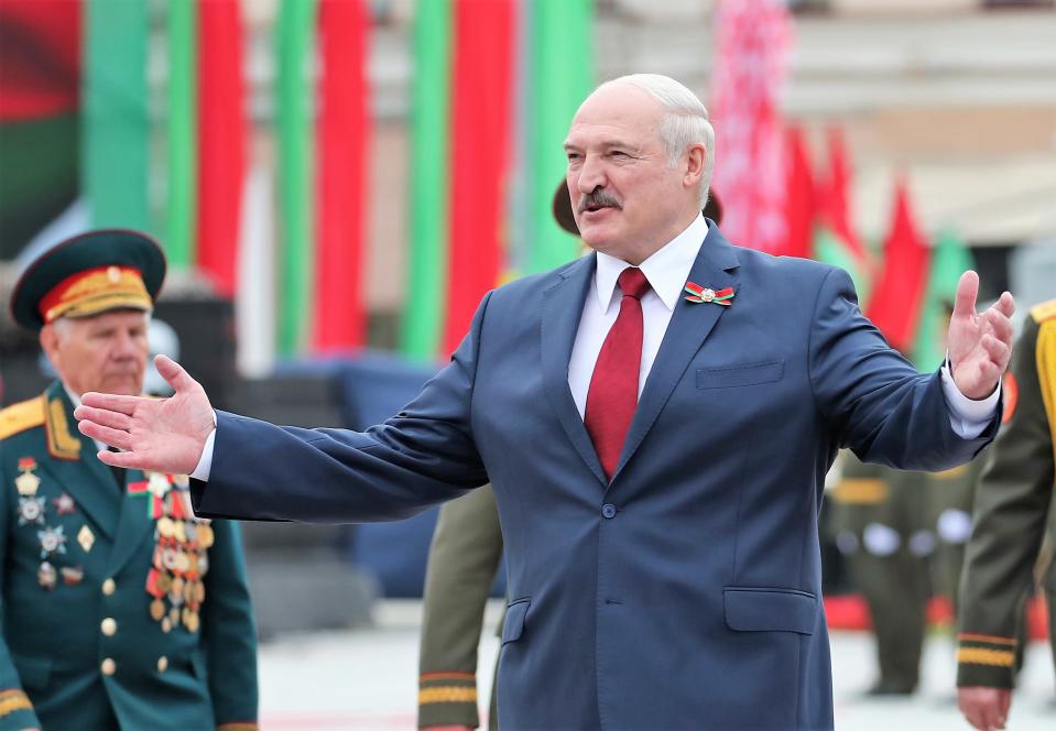 Lukashenka levittää käsiään hymyillen. Hänellä on tumma puku, punainen kravatti. Taustalla seisoo vanha mies univormussa.