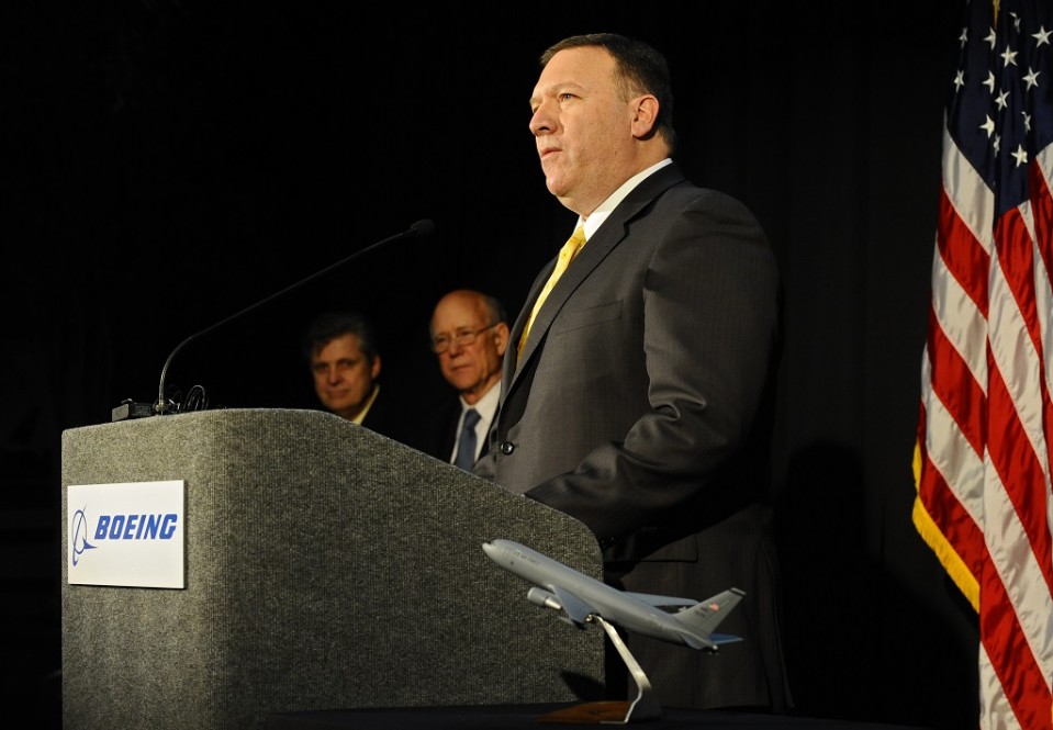 Mike Pompeo seisoo puhujankorokkeen ääressä tummassa puvussa, keltaisessa kravatissa ja katsoo vakavana yleisöön. Taustalla on Yhdysvaltain lippu ja muita henkilöitä.