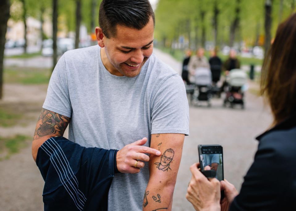 Mies näyttää käsivarrestaan tatuointia kännykkäkameralla kuvaavalle naiselle.