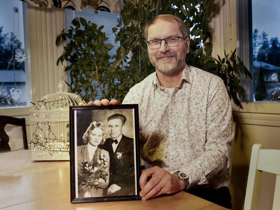 Mika Koivunen istuu pöydän äärellä, katsoo kameraan hymyillen ja pitelee vanhempiensa hääkuvaa.