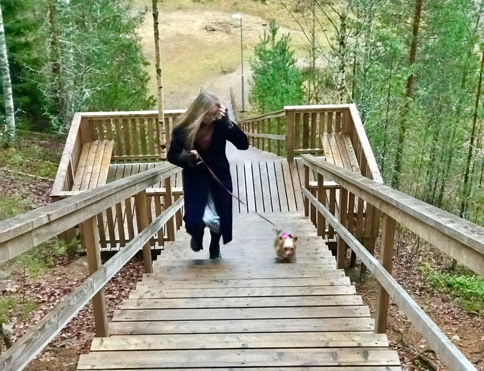 Vaalea, pitkätukkainen nainen lilassa pitkässä takissa juoksee ylös kuntoportaita pienen koiran kanssa