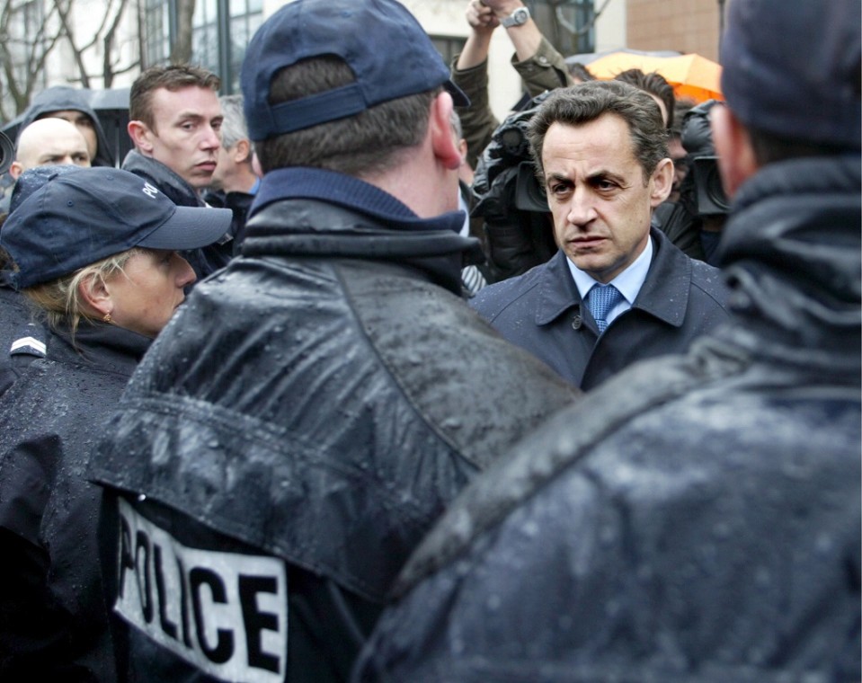 Ryhmä poliiseja on selin kameraan. Heidän takeillaan on sadepisaroita. Sarkozy seisoo heidän edessään vakavan näköisenä tummassa päällystakissa, sininen kravatti kaulassaan.