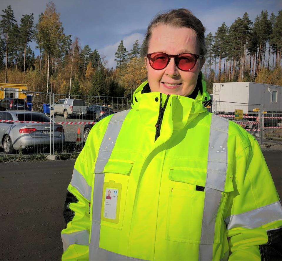 Huomiovaatteissa oleva nainen hymyilee auringossa kuvaajalle