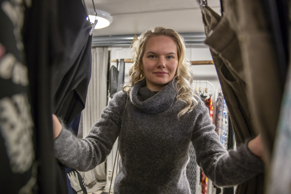 Nainen katsoo kameraan vaaterekissä olevien vaatteiden takaa.