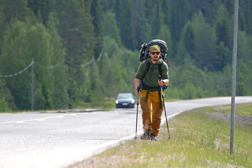 Mies kävelee valtatien reunaa pitkin rinkka selässään.