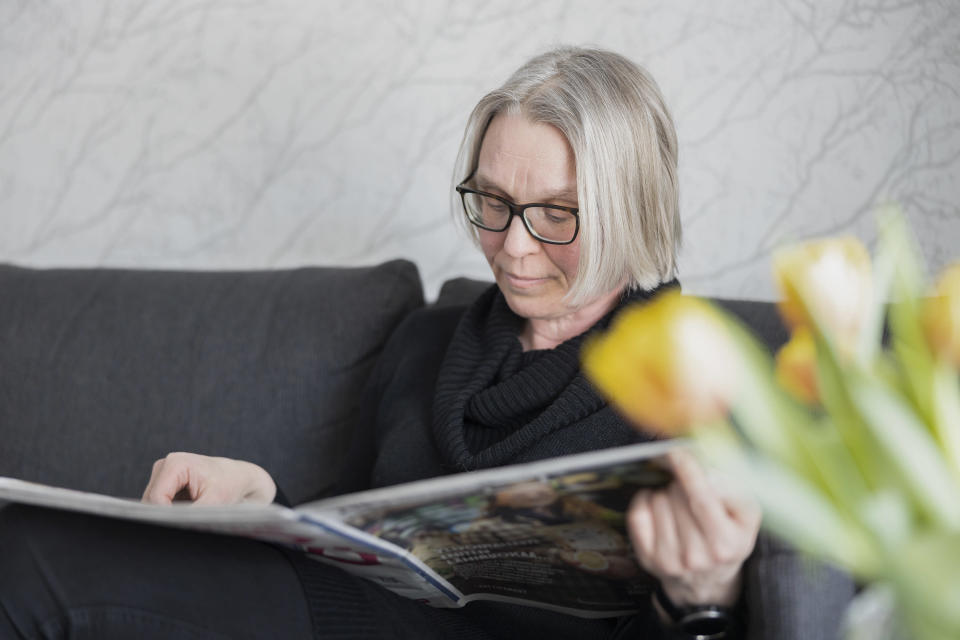 Anne Matilainen istuu olohuoneen sohvalla ja lukee sanomalehteä. Kuvan etualalla on keltaisia tulppaaneja.