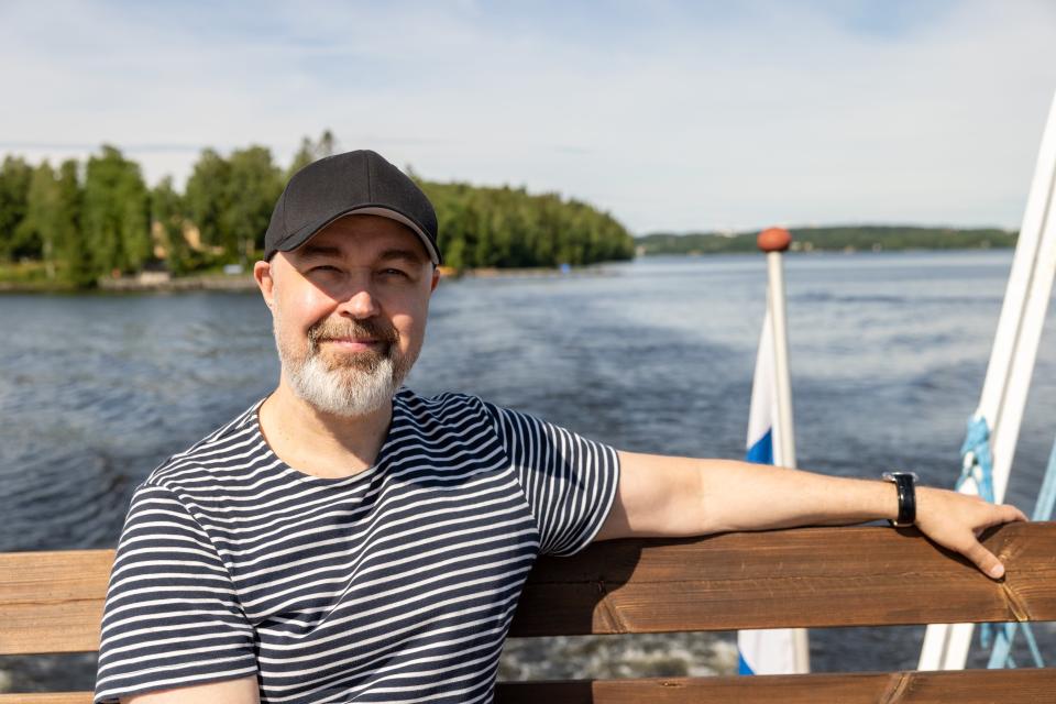 Harmaapartainen mies mustassa lippalakissa ja mustavalkoraidallisessa t-paidassa katsoo kameraan hymyillen. Mies istuu laivan kannella nojaten kaiteeseen, taustalla näkyy aurinkoista Pyhäjärveä.