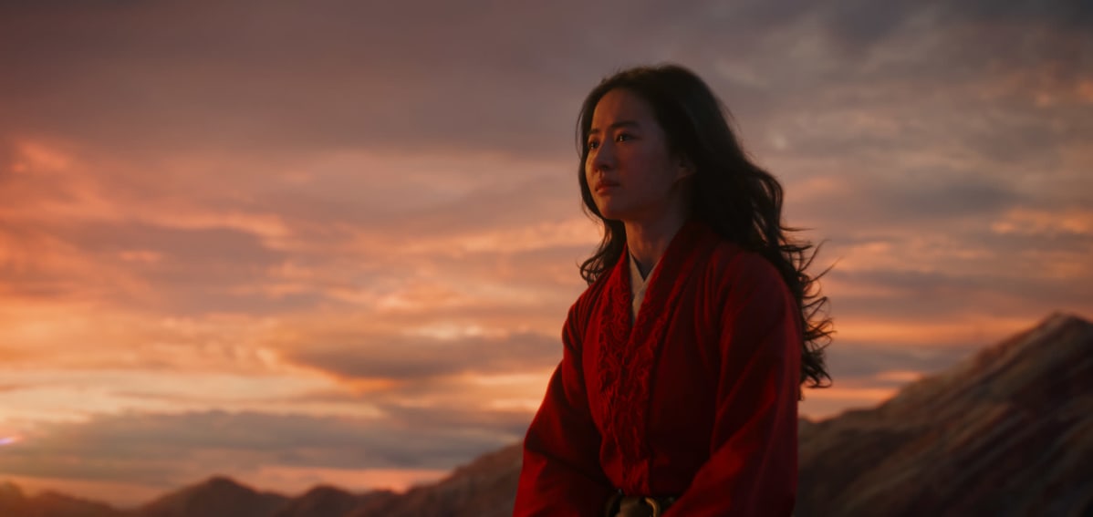 Närbild på Mulan (Yifei Liu) sitter på ett berg i solnedgången.