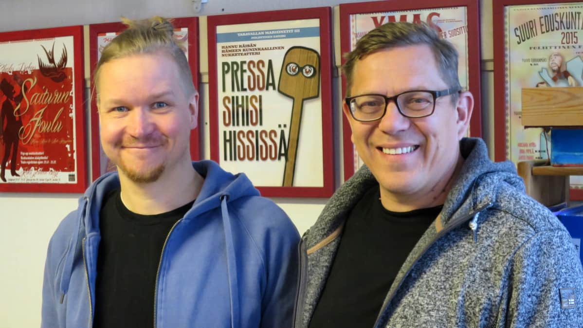 Kaksi miestä hymyilee julisteiden edessä