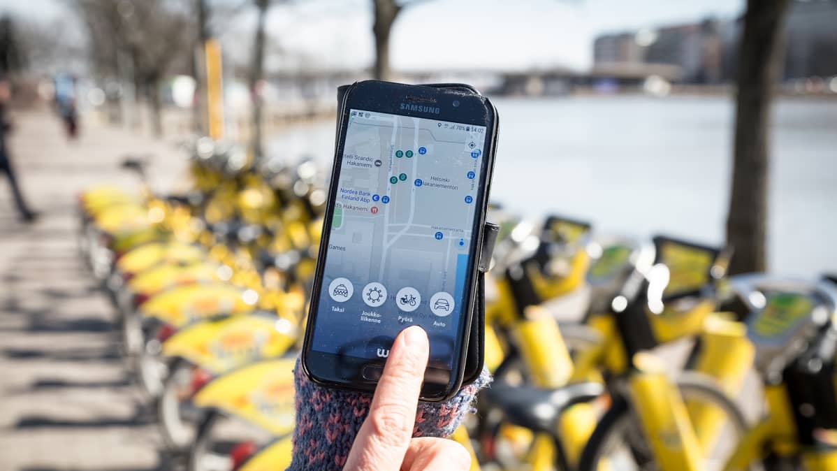 Whim-mobiilisovellus älypuhelimen näytöllä, taustalla kaupunkipyöriä.