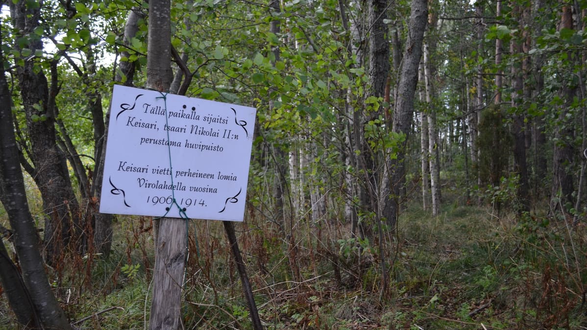 Valkoinen kyltti metsässä. Kyltissä kerrotaan paikalla sijainneen keisari, tsaari Nikolai II:n perustama huvipuisto.