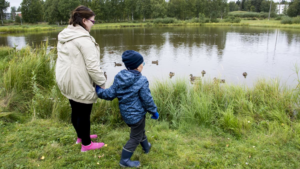 Äiti on pienen poikansa kanssa ruokkimassa sorsia lammen rannalla.