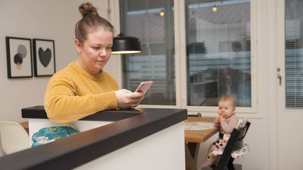 Äiti katsoo älypuhelinta ja vauva istuu syöttötuolissa taka-alalla.