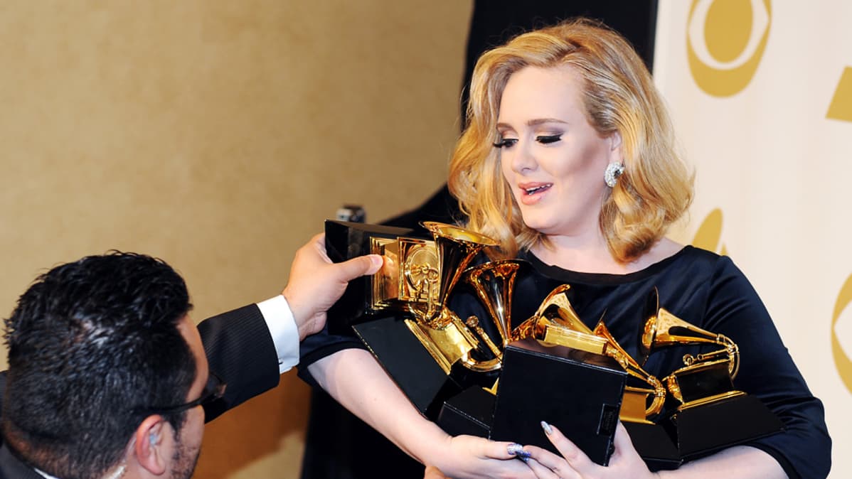 Avustaja asettelee Grammy-palkintoja Adelen syliin.