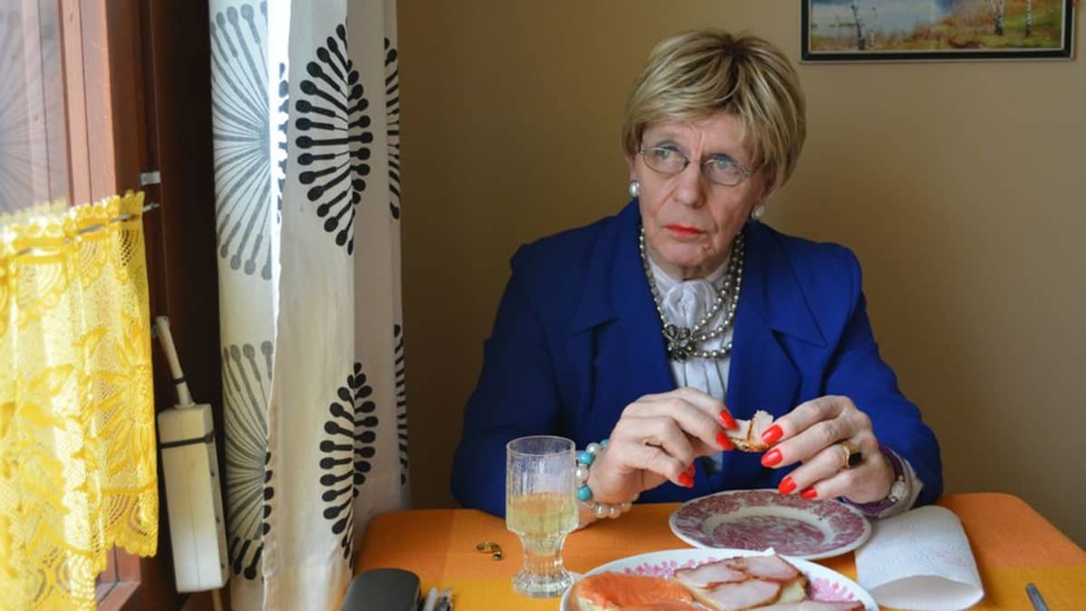 Mieheksi pukeutunut nainen istuu keittiönpöydän ääressä ja syö voileipää.