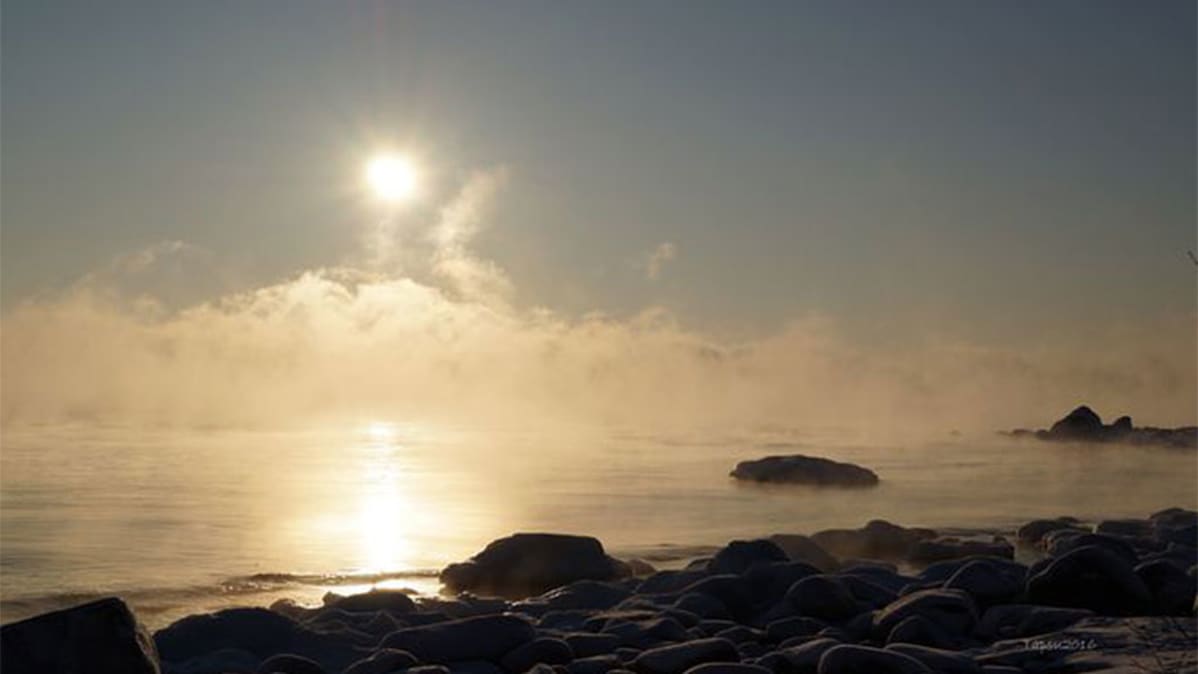 Mereltä, etualalla kivistä rantaa jää päätyy sumuun, aurinko kaiken yllä.
