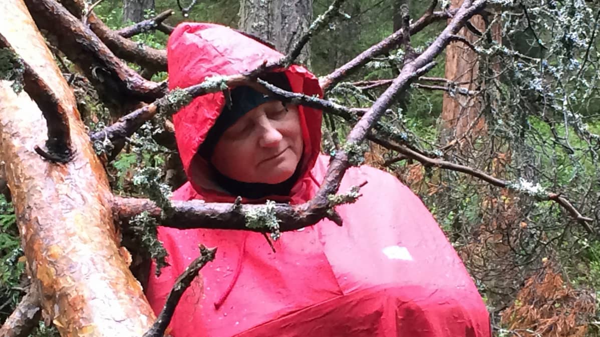 Punaiseen sadetakkiin pukeutunut nainen seisoo metsässä silmät kiinni.