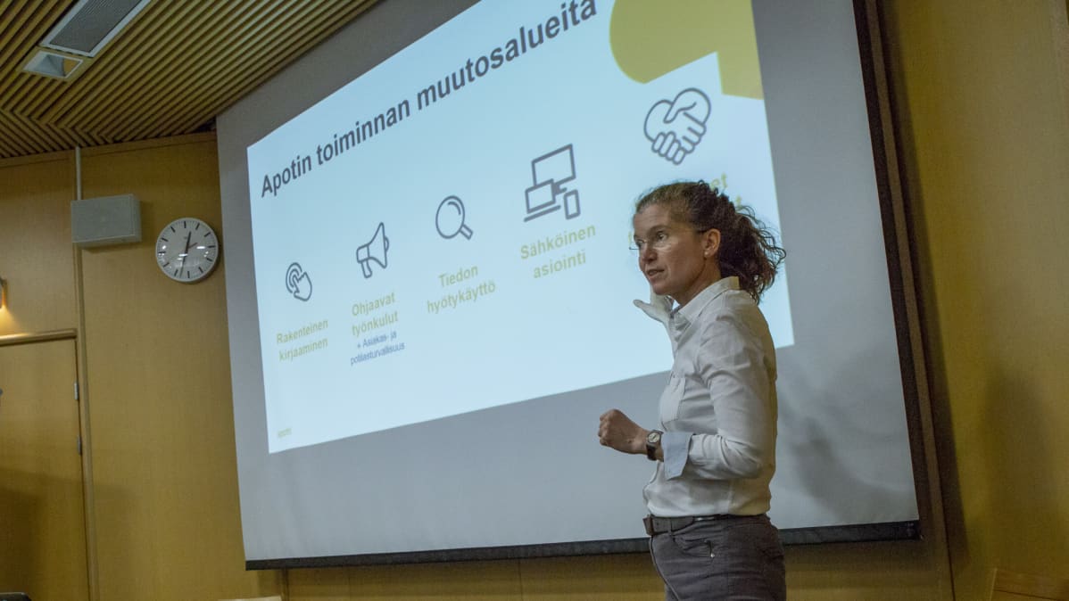 En kvinna, Piia Vuorela, står och pekar mot en powerpoint presentation om den nya patientinformationssystemet Apotti.