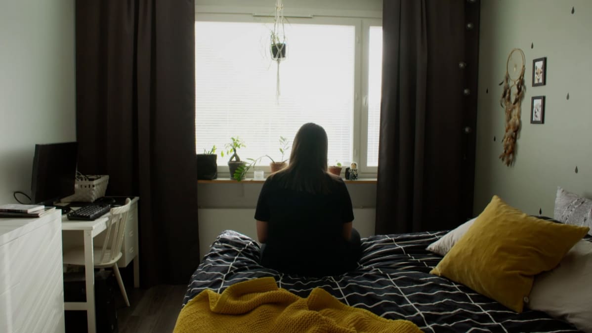 Nainen istuu sängyn reunalla ja katsoo ikkunasta ulos.